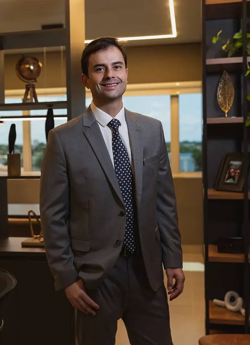 Retrato profissional do Dr. Rodrigo Marçal apoiado em cadeira de design metálico, vestindo camisa social azul clara, expressão serena e contemplativa olhando para o lado, fundo escuro neutro em tom carvão, iluminação lateral suave e dramática estilo estúdio, composição vertical sóbria, fotografia editorial ultra-realista em alta definição