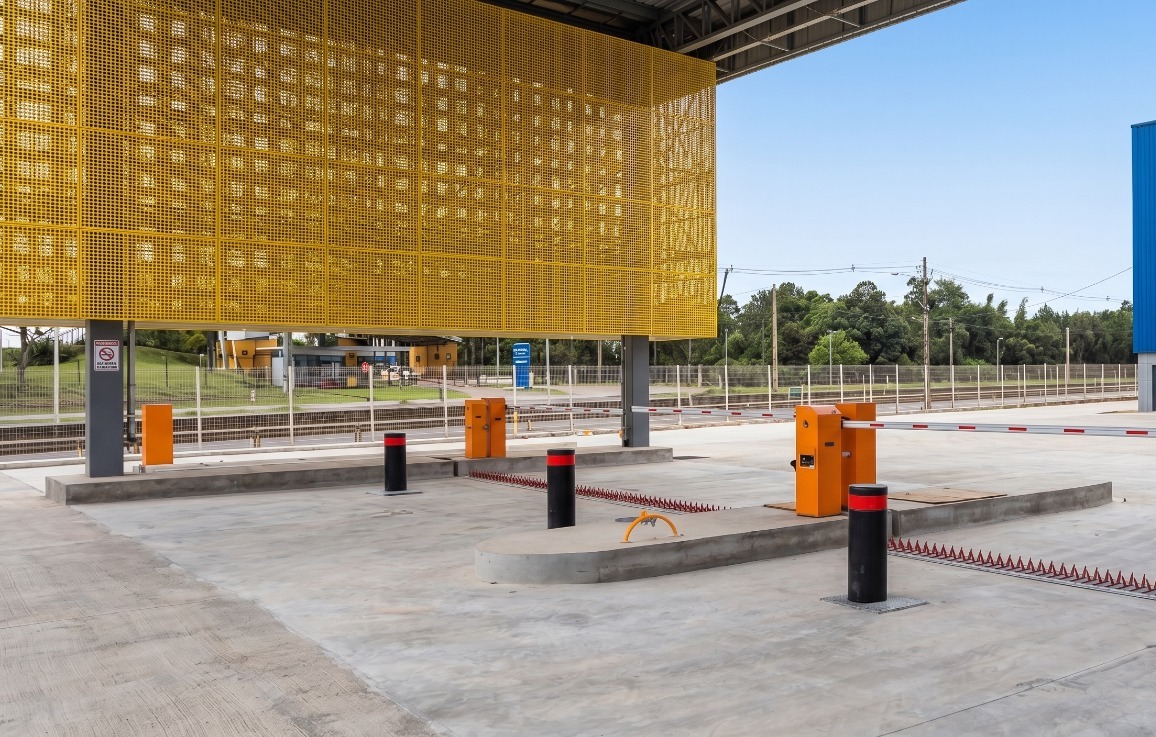 Portaria veicular com fachada amarela perfurada decorativa, dois bollards automáticos pretos com faixa refletiva vermelha em ilha central de concreto, cancelas laranjas e spike barrier vermelho ao chão, vista lateral em ambiente externo diurno, fotografia industrial ultra-realista
