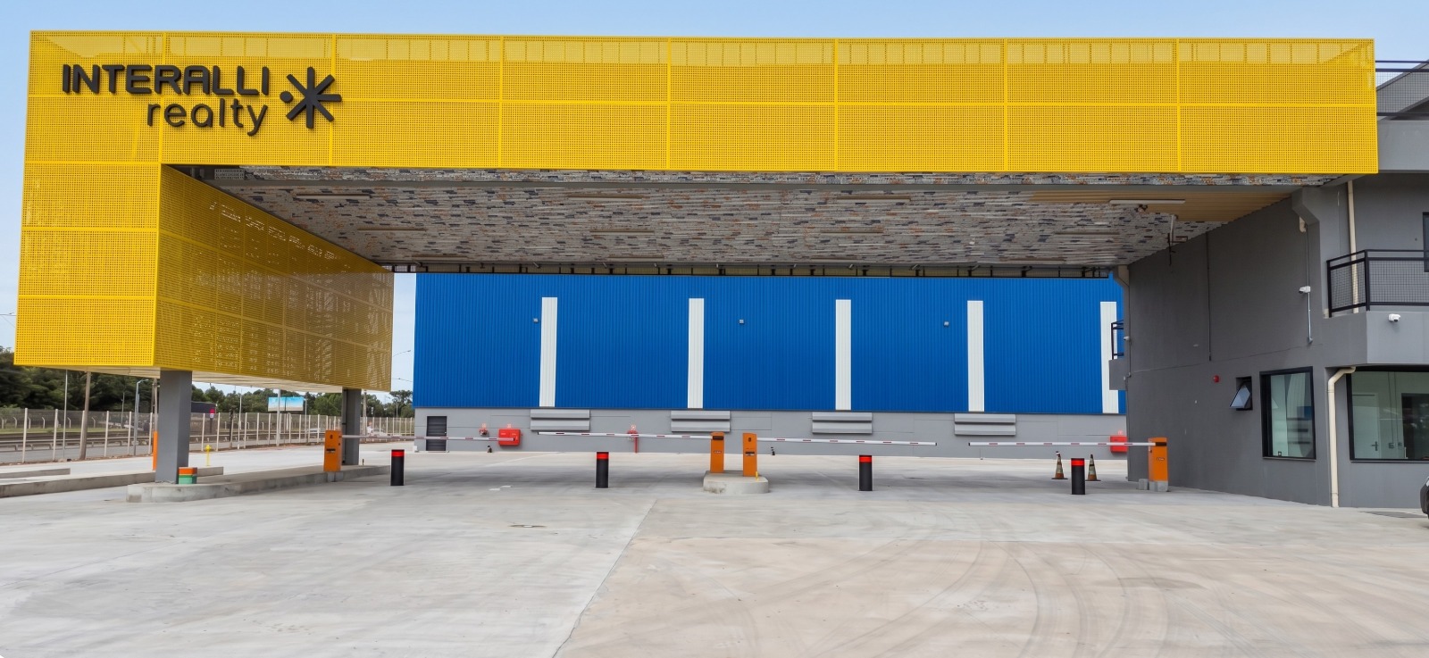 Portaria de centro logístico Interalli Realty com fachada amarela perfurada, eclusa de segurança com bollards automáticos pretos com faixa refletiva vermelha, cancelas laranjas, pavimento de concreto e galpão azul ao fundo, fotografia industrial em alta resolução com luz natural diurna