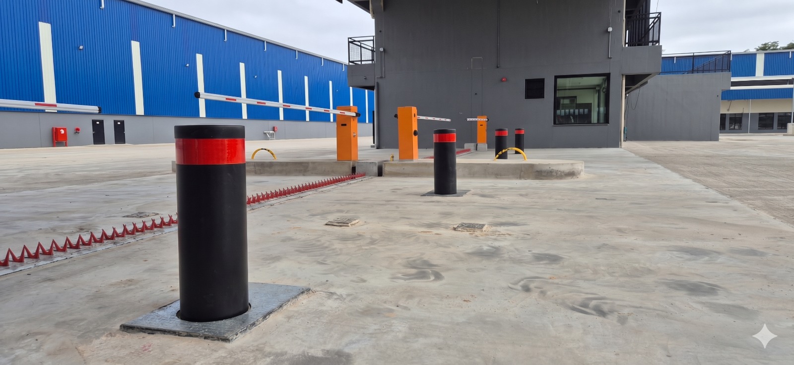 Bollards automáticos pretos com faixa refletiva vermelha instalados em pavimento de concreto industrial, spike barrier vermelho ao chão, cancelas laranjas e galpão de operação logística azul ao fundo, fotografia ultra-realista de instalação de segurança em ângulo baixo dramático